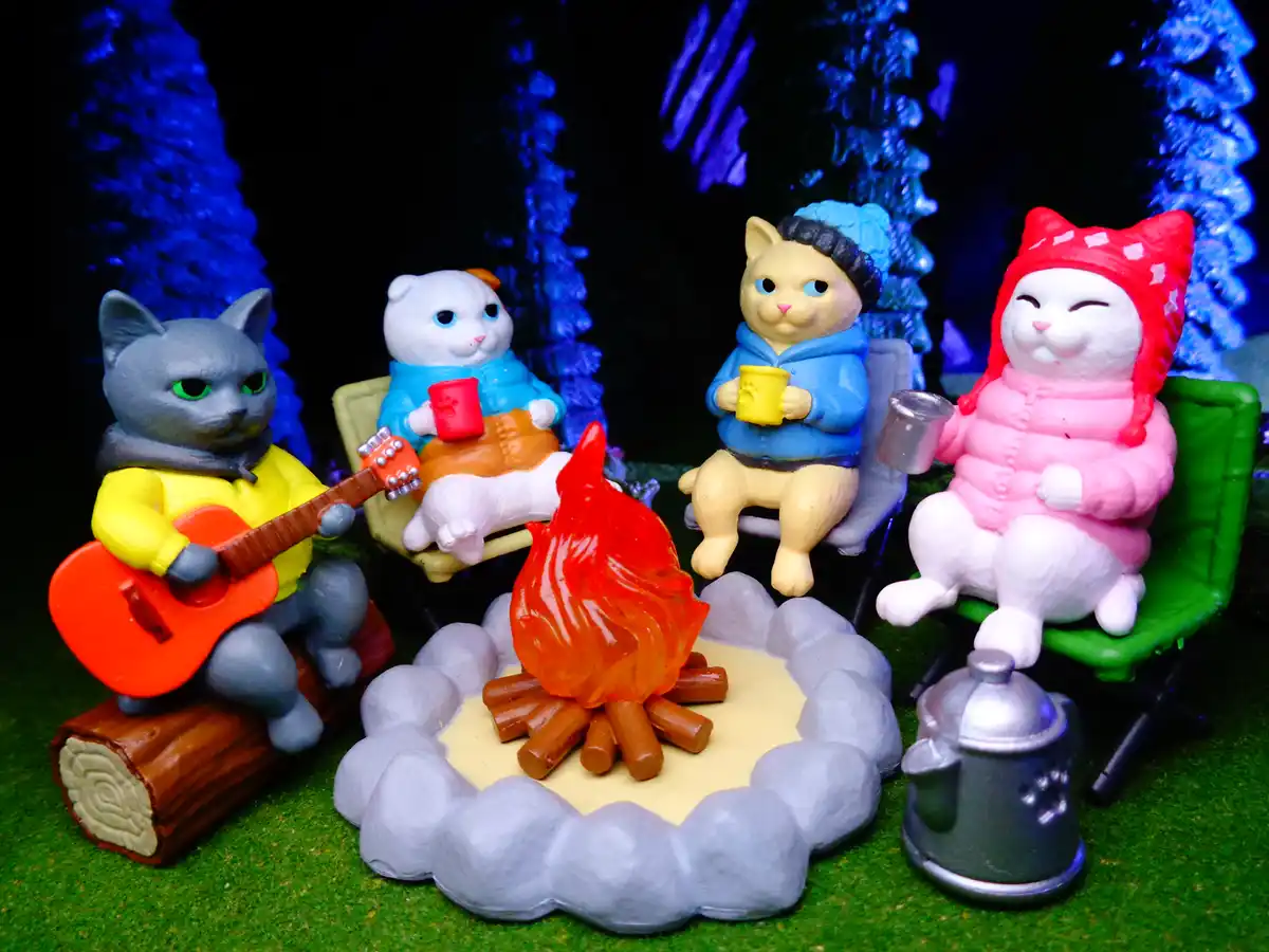 Four plastic cat figurines are gathered around a miniature campfire in a forest setting, each wearing cozy winter clothes…