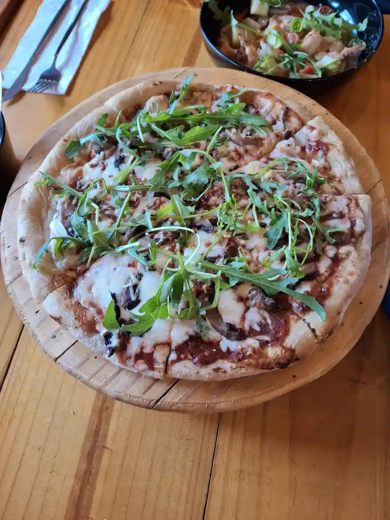 A freshly baked pizza topped with melted cheese, mushrooms, and a generous amount of fresh arugula sits on a wooden serving…