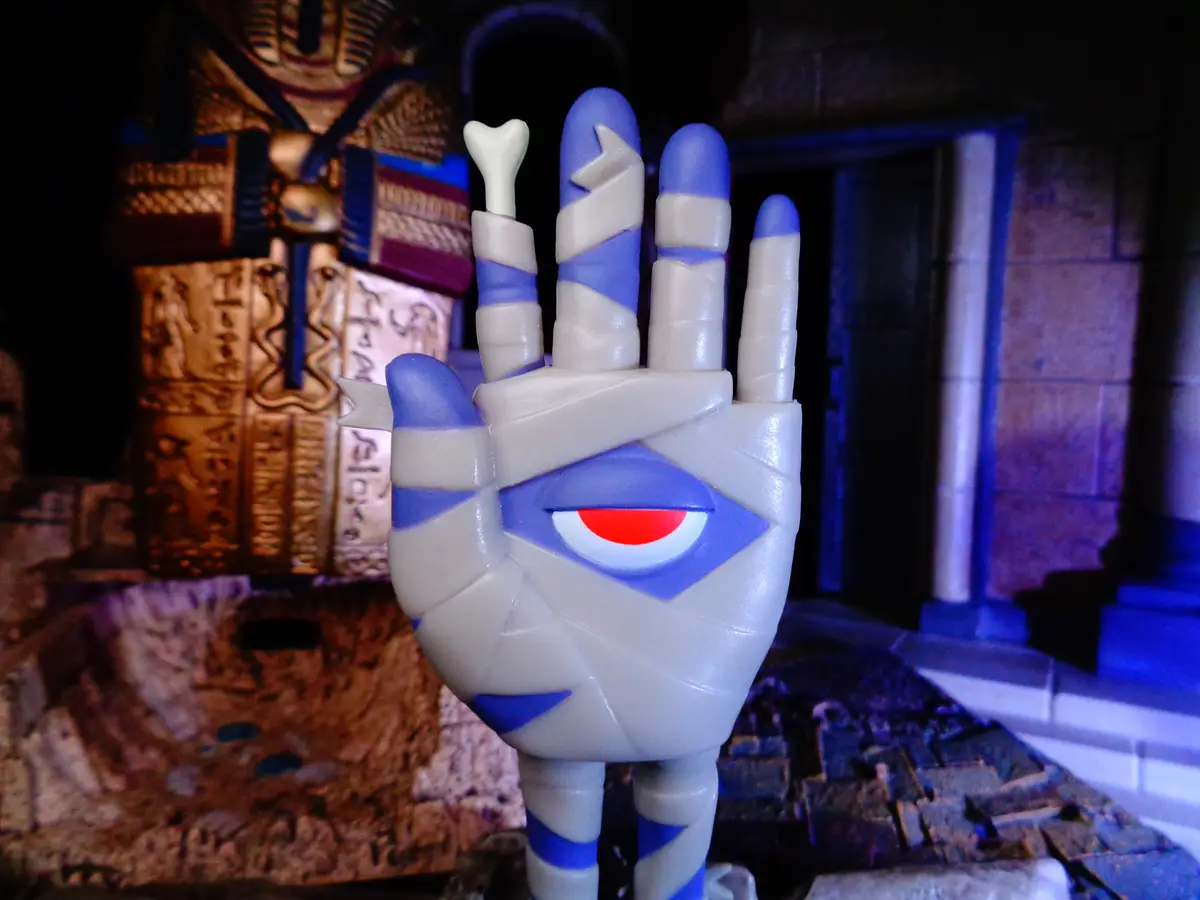 A close-up of a stylized mummy hand statue with blue and white wrappings, featuring a glowing red eye in the palm and a…
