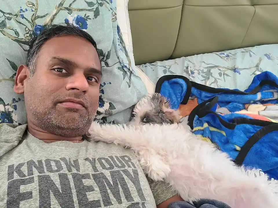 A man with gray-streaked hair lies on a floral-patterned couch, wearing a gray t-shirt that reads 'KNOW YOUR ENEMY'. A…