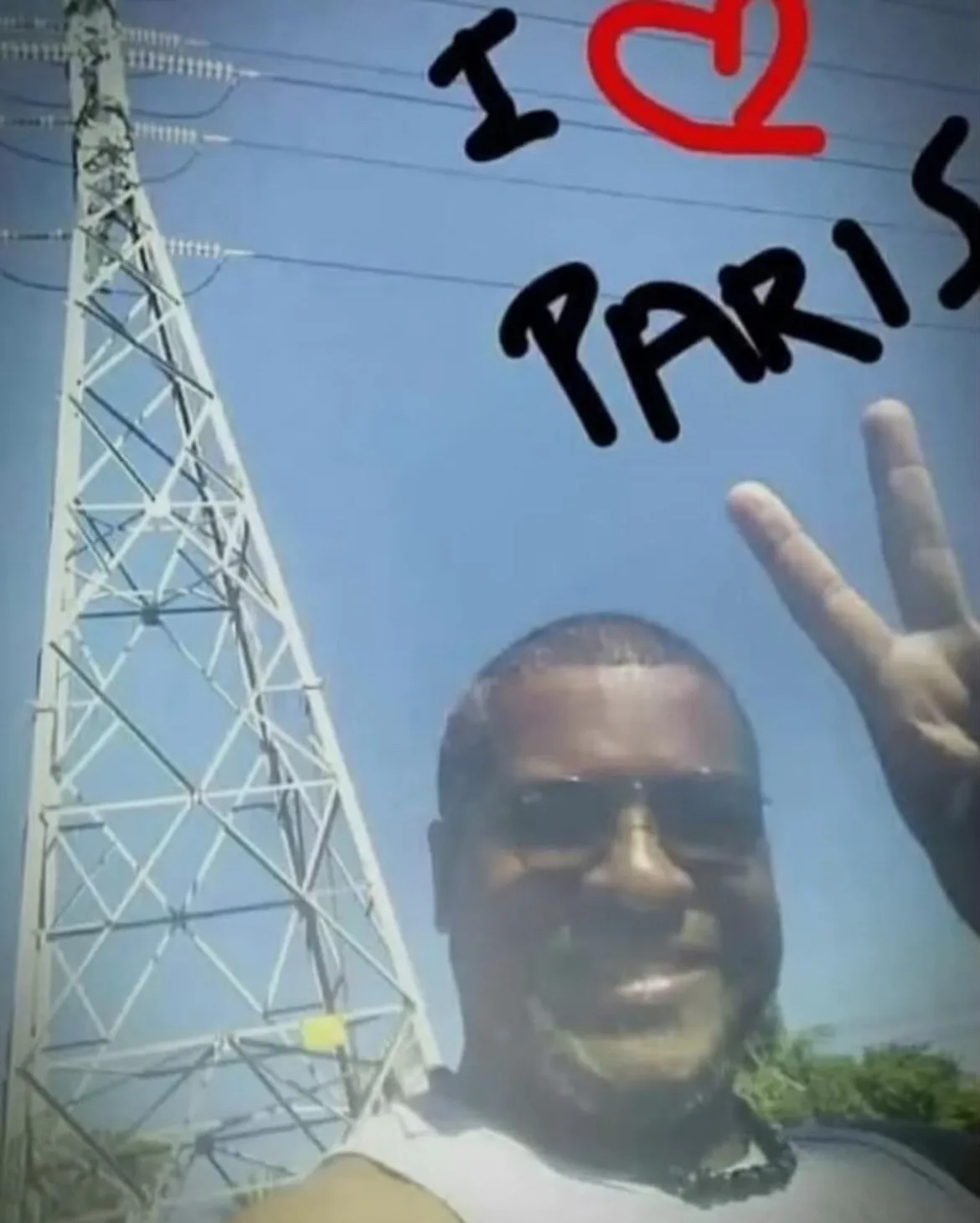 A man wearing sunglasses and a white tank top smiles at the camera while holding up a peace sign, standing in front of a…