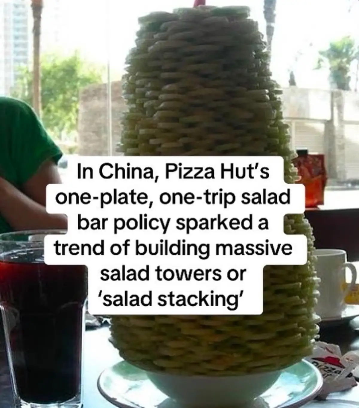 A towering, cone-shaped stack of green salad ingredients sits on a white plate, resembling a precarious culinary structure…