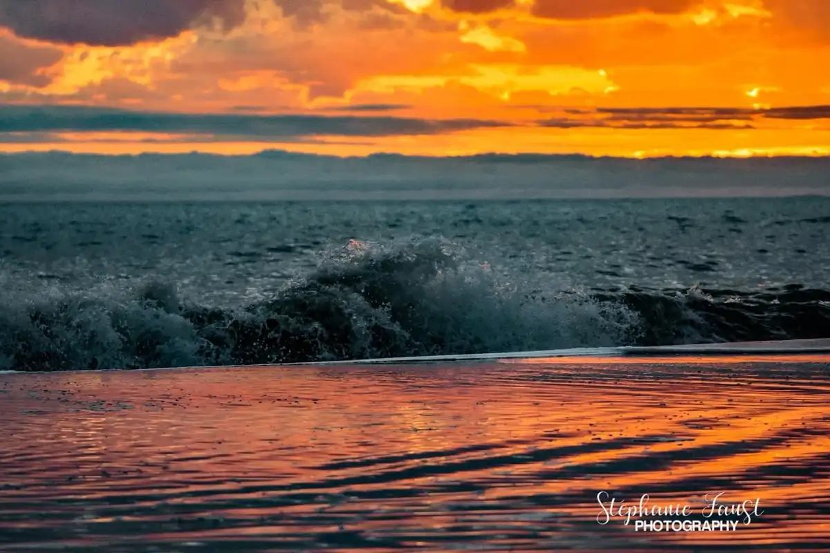 A serene sunset over the ocean with vibrant orange and yellow clouds reflecting on the water's surface. A wave crashes in…