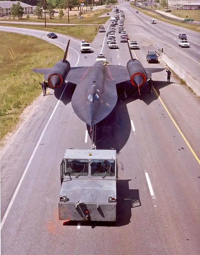 An aerial view shows a black, stealthy SR-71 Blackbird reconnaissance aircraft being towed by a small military truck down a…