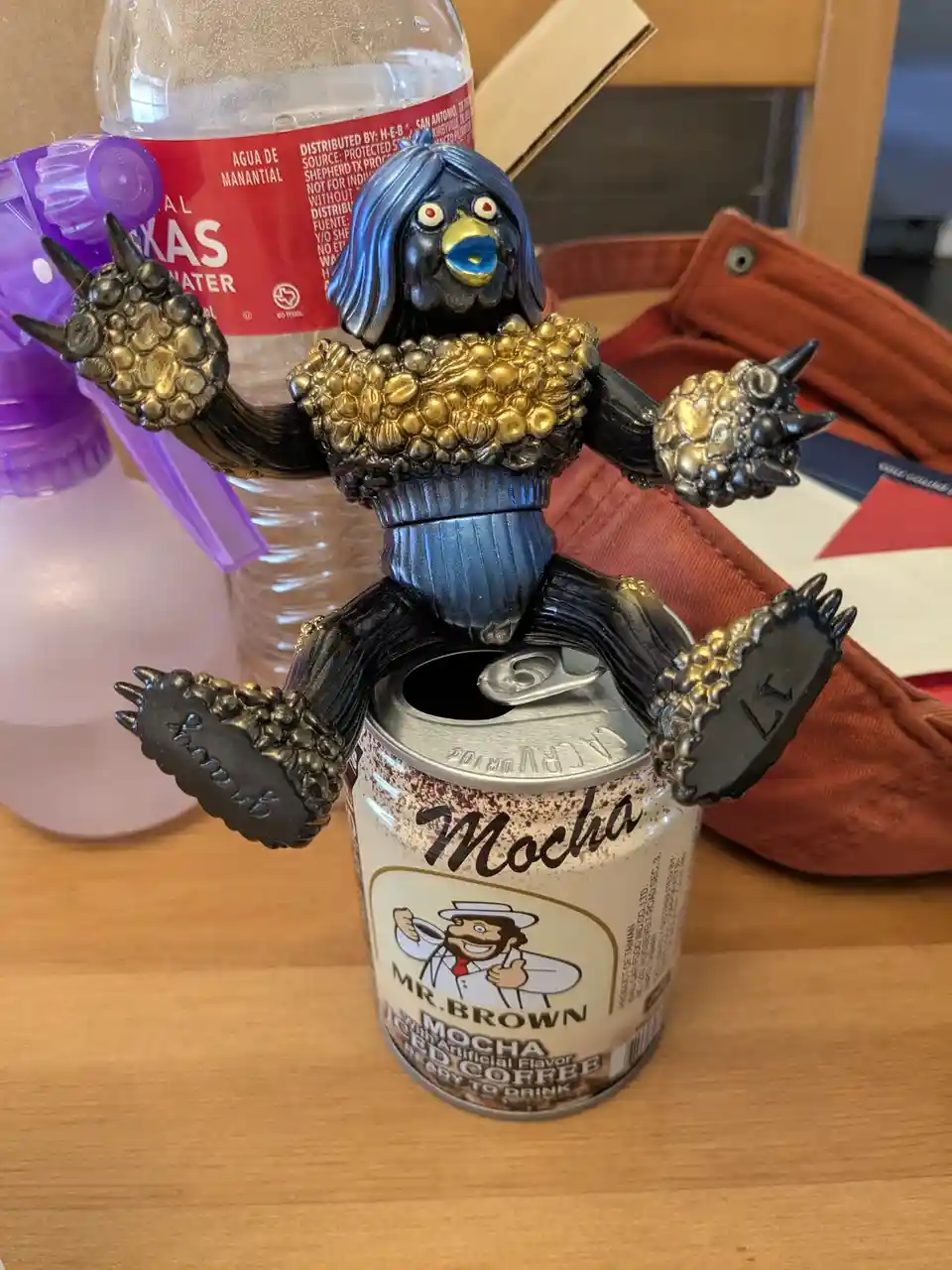 A black, bird-like figurine with blue hair and gold accents sits atop an open can of Mr. Brown Mocha iced coffee, its clawed…