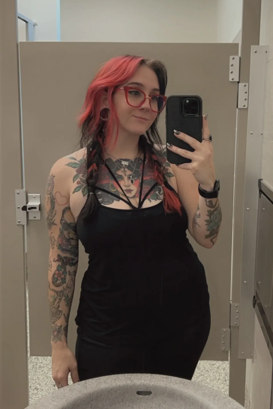 A person with red and black hair, red glasses, and extensive tattoos on their arms and chest takes a mirror selfie in a…