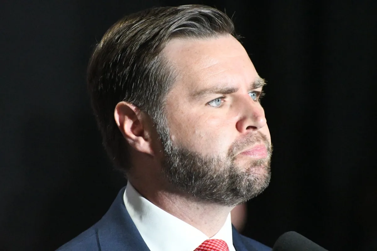 A close-up photo of a man with a beard and blue eyes, wearing a suit and red tie, looking off to the side with a serious…
