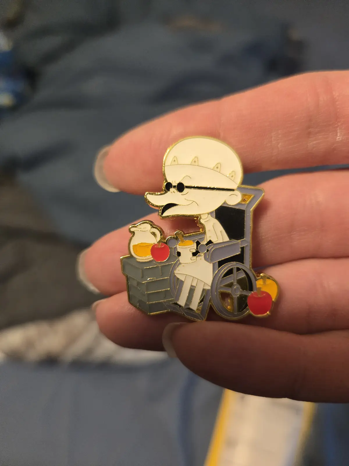 A hand holds a metal pin featuring the cartoon character Mr. Pickles from 'The Simpsons' in a wheelchair, wearing sunglasses…