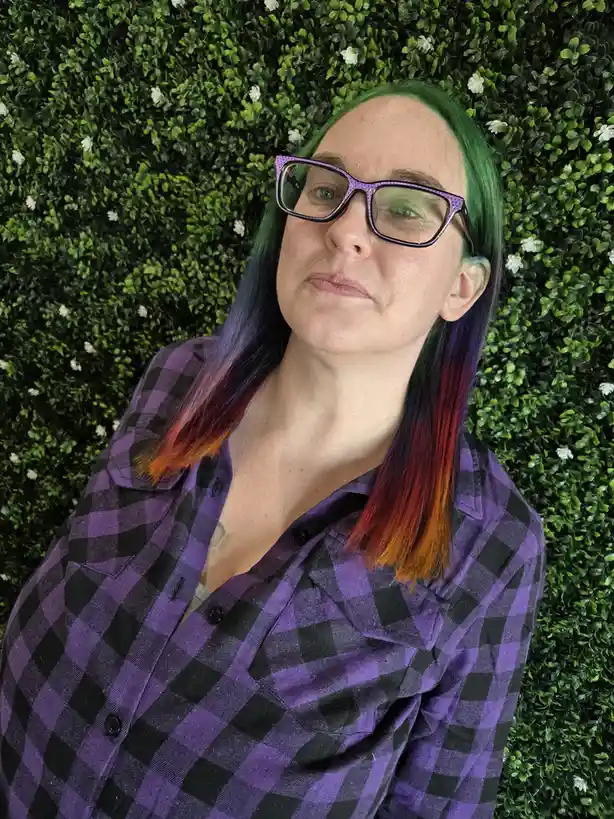 A person with vibrant green-to-purple-to-orange-red ombre hair and purple polka-dot glasses wears a purple and black plaid…