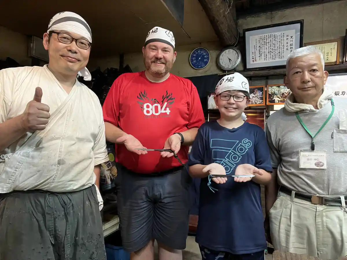 Four people, including a boy and two men in white headbands, stand in a workshop holding small knives. The man on the left…