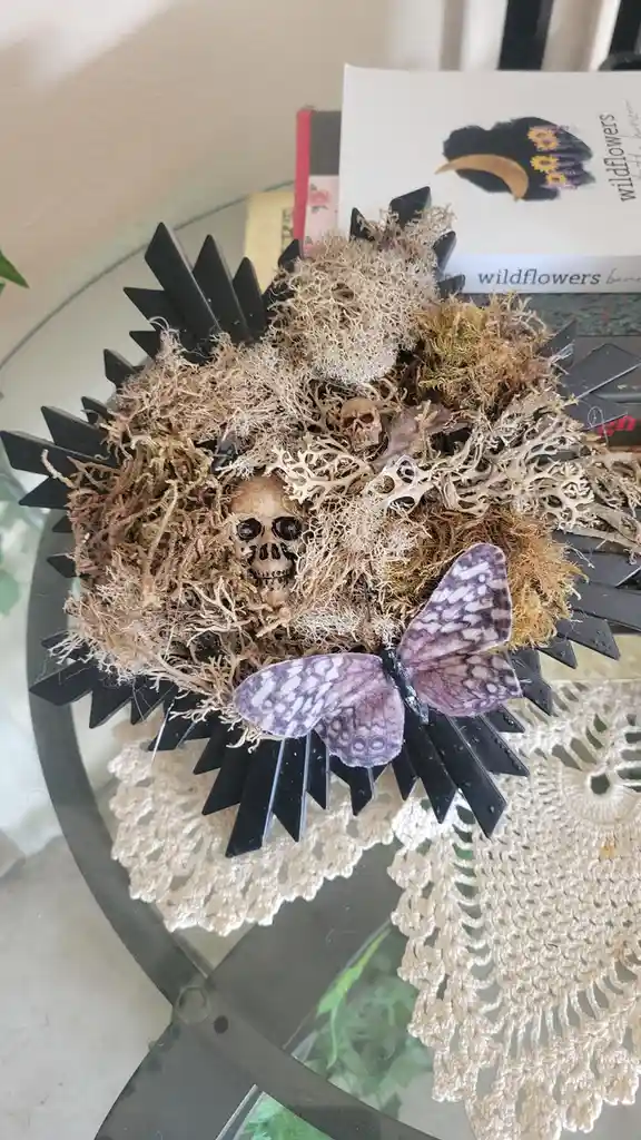 A decorative wreath made of dried moss, small skulls, and black spiked elements sits on a glass table with a lace doily. A…