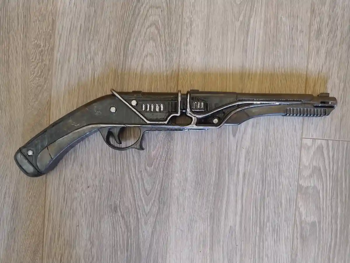 A dark, metallic toy gun with a handle lies on a light wood-grain floor. The overlaid text reads 'i'm not a gun, i'm a gun…