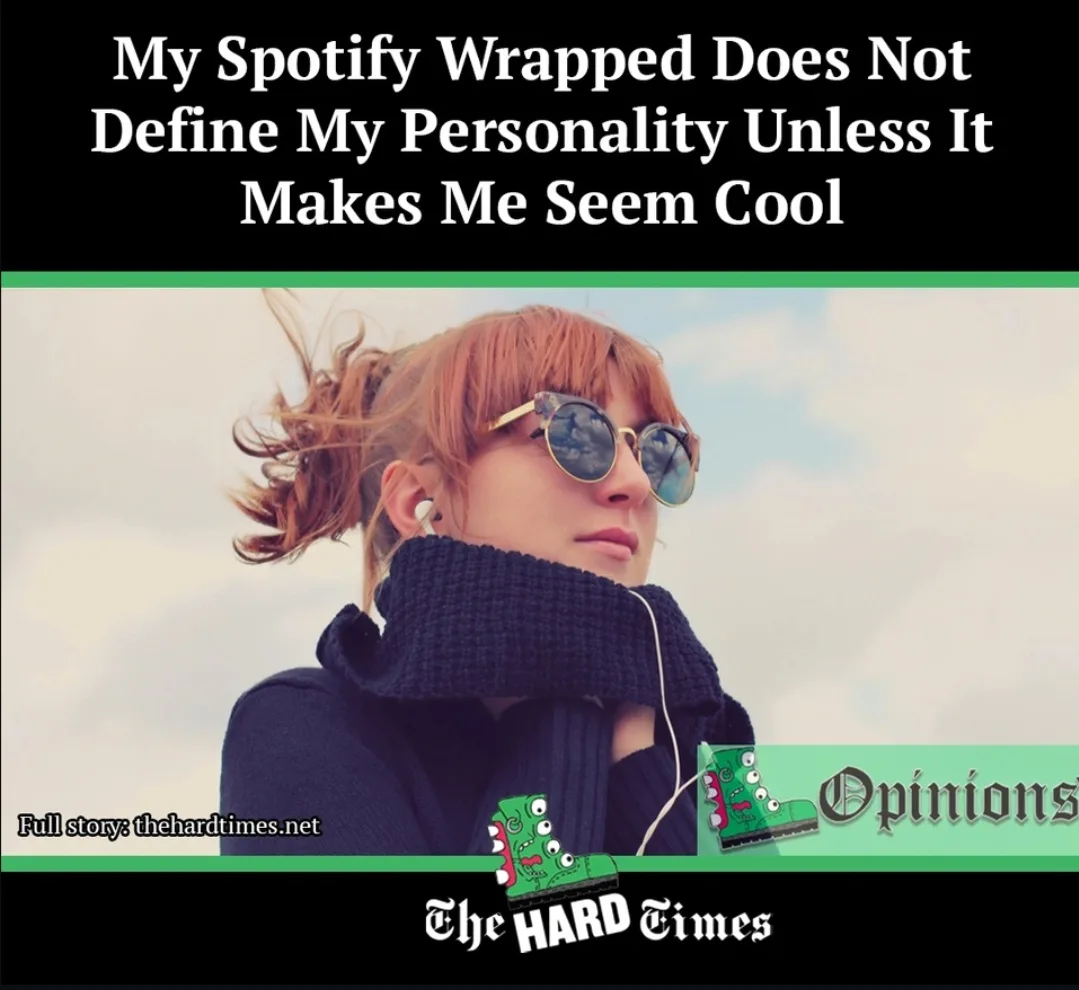 A woman with reddish hair in a ponytail, wearing round sunglasses and a thick dark blue scarf, listens to music via white…
