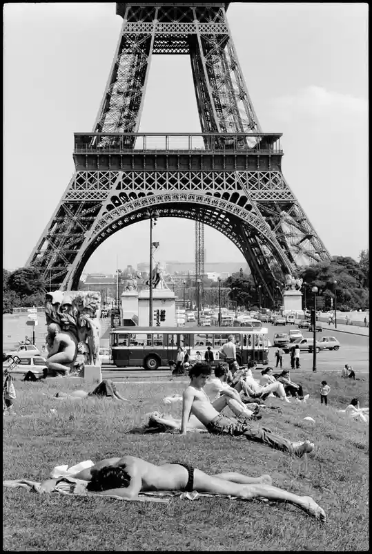 A black-and-white photo shows people sunbathing on grass in front of the Eiffel Tower, with a bus and cars visible in the…