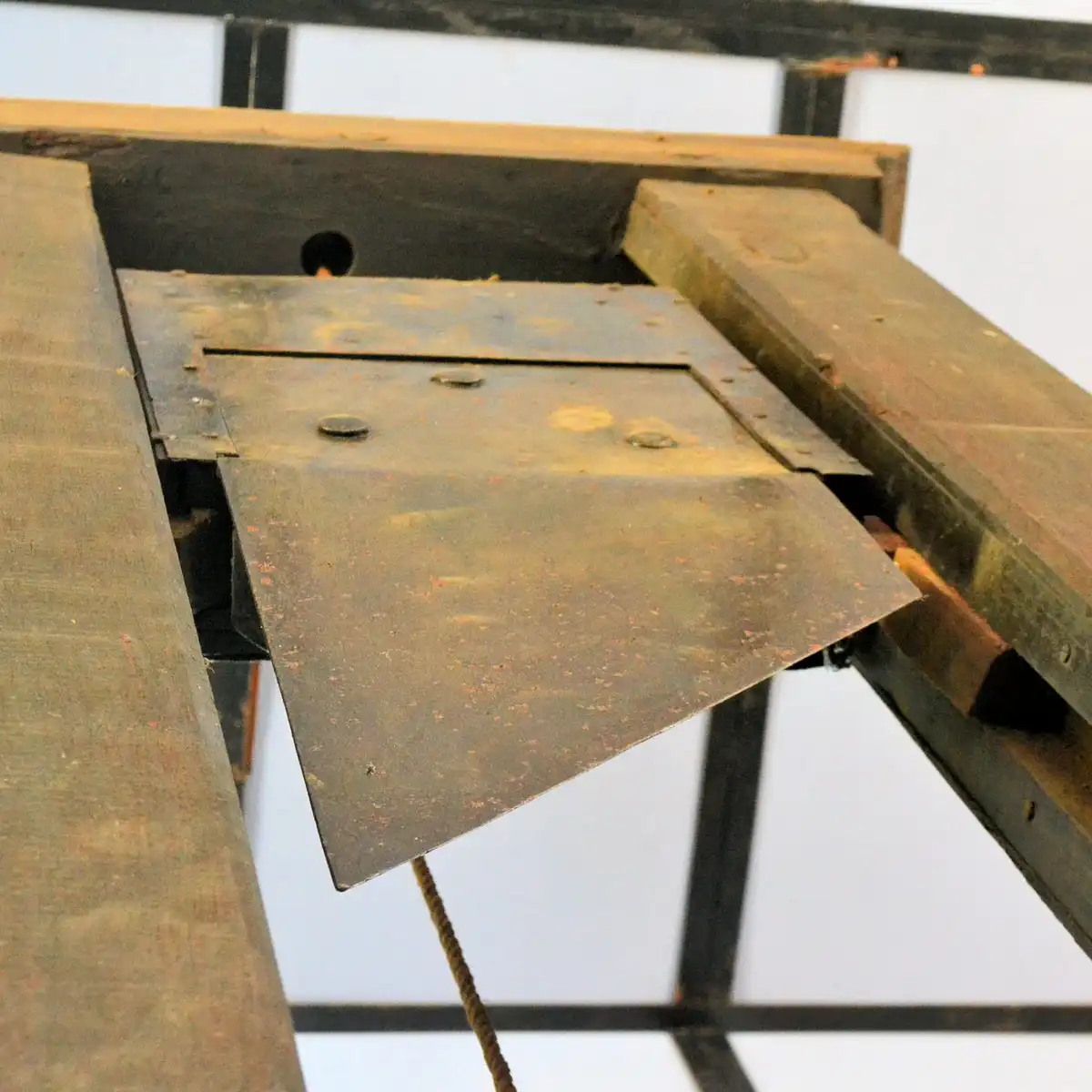 A close-up of a rusty, hinged metal trapdoor on a wooden structure, partially open, revealing a dark cavity beneath. The…