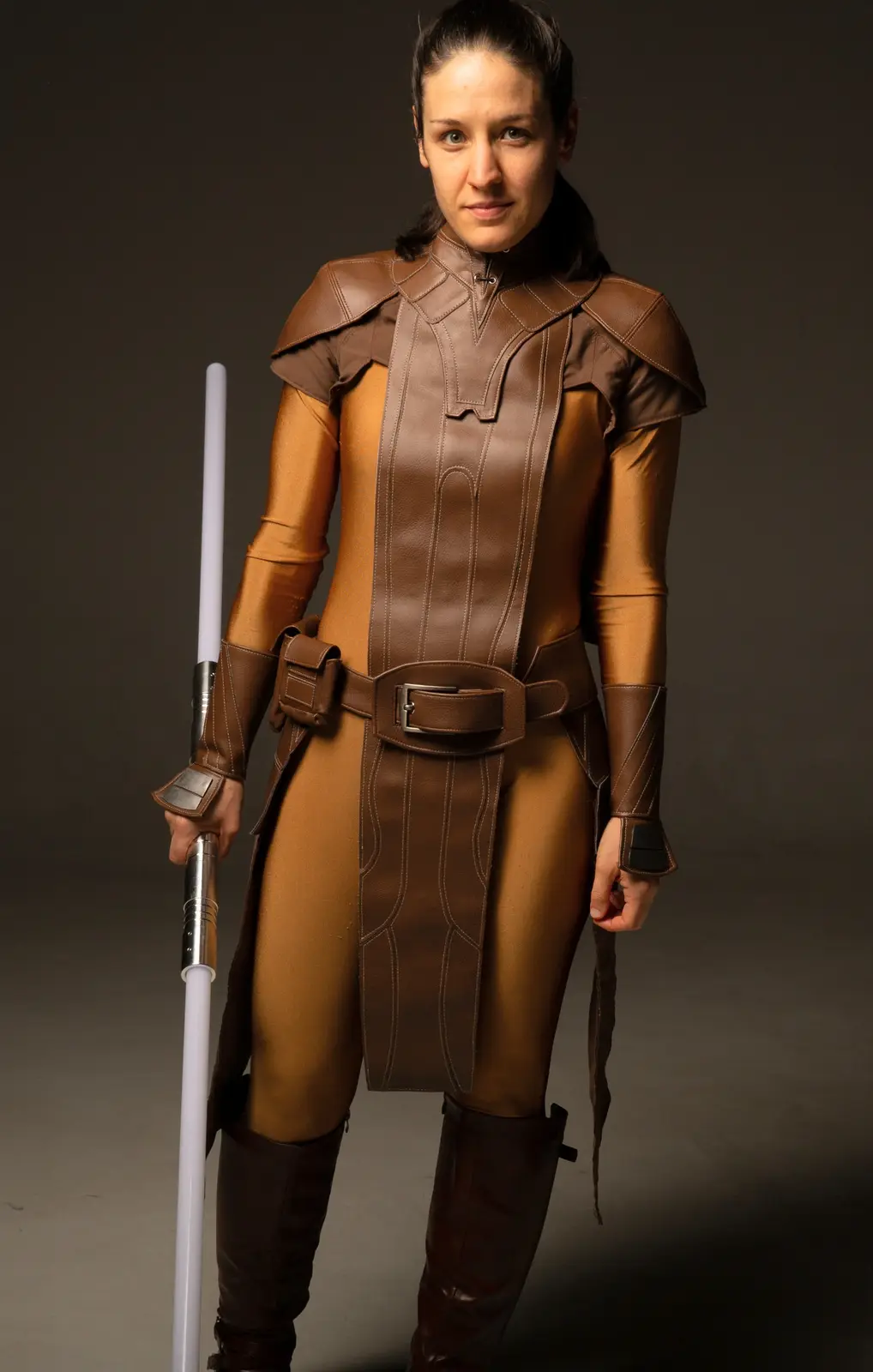 A woman in a brown and gold Jedi-style costume holds a white lightsaber, looking directly at the camera with a neutral…