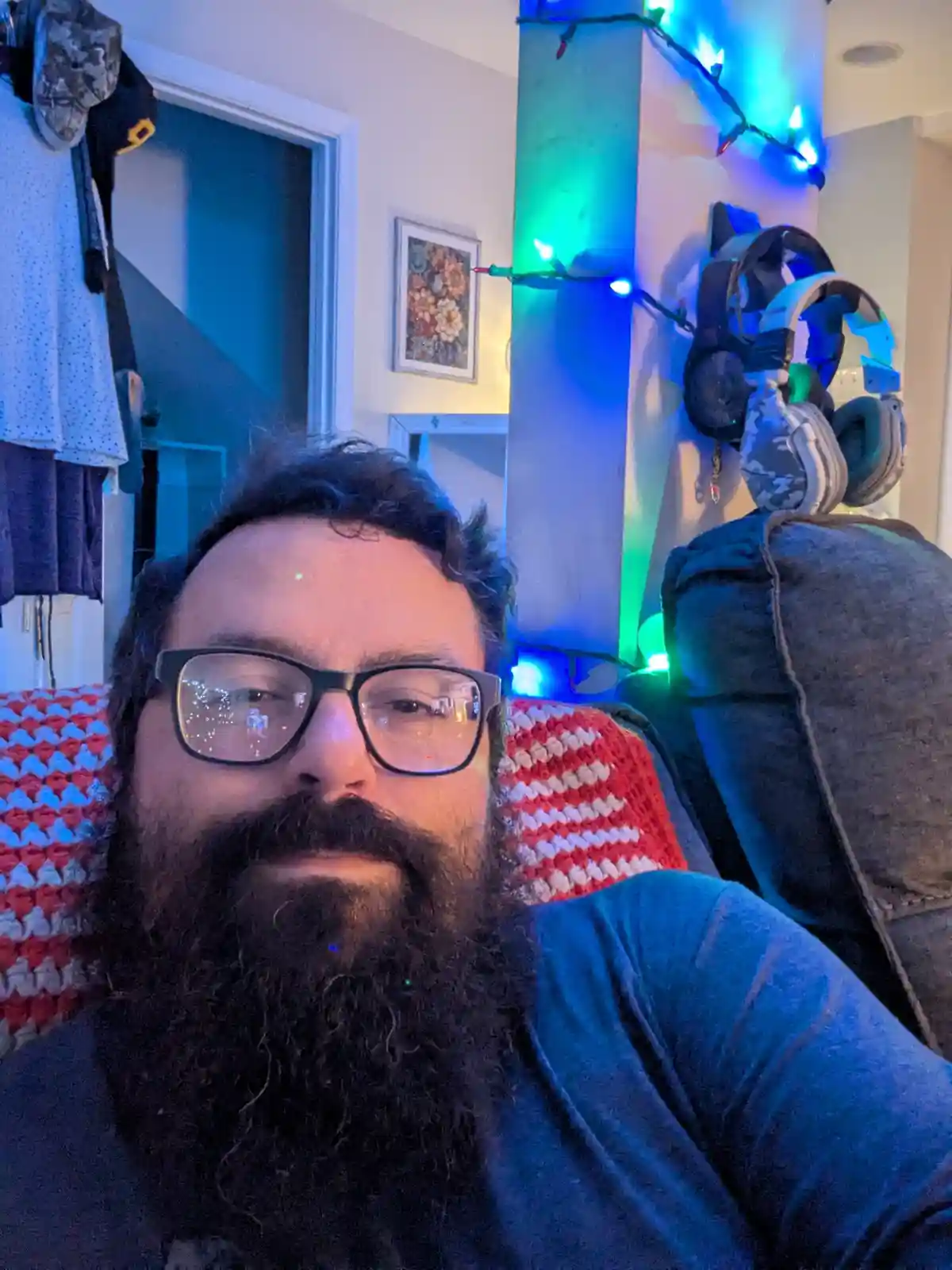 A man with a full beard and glasses sits on a couch with a red and white patterned blanket, surrounded by colorful string…