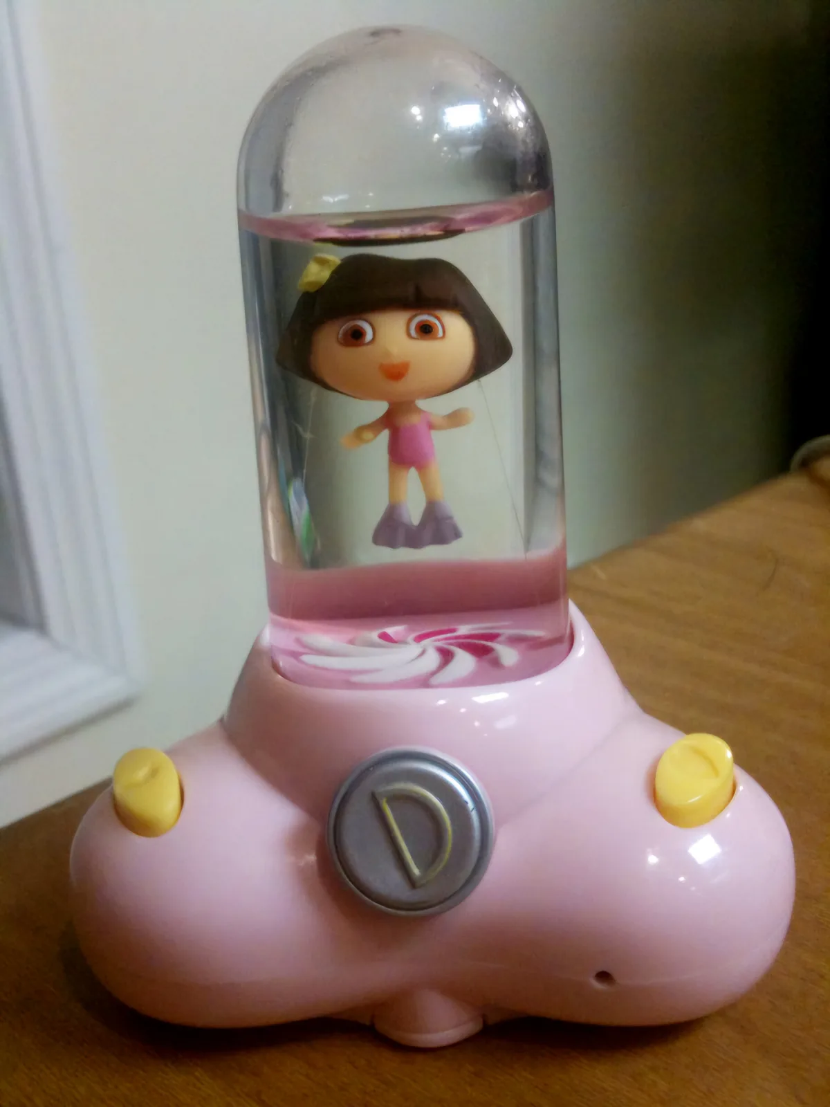 A pink, car-shaped children's toy with a clear dome containing a Dora the Explorer figurine floating in pink liquid. The…