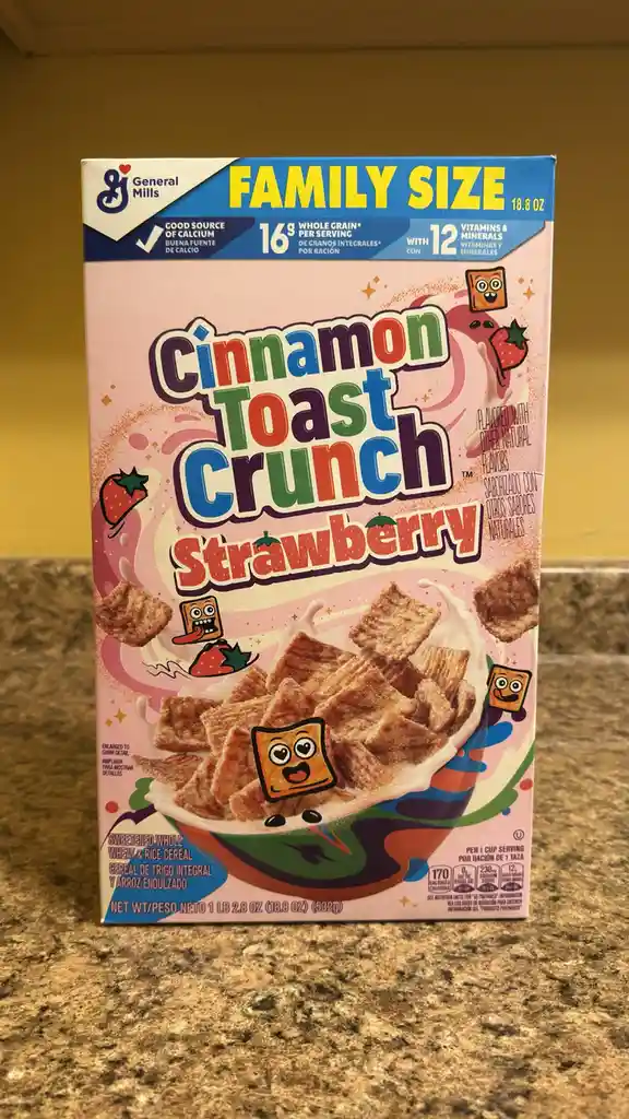A family-size box of General Mills Cinnamon Toast Crunch Strawberry cereal sits on a granite countertop. The box features…