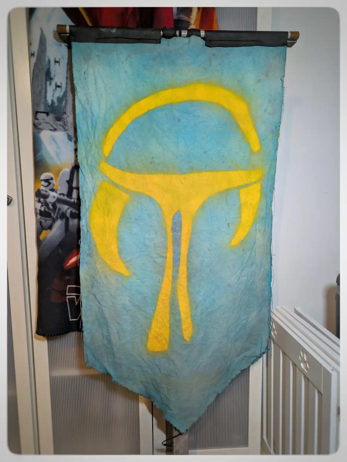 A handmade, light blue fabric banner with a yellow Mandalorian helmet symbol hangs on a black rod in a room. To the left, a…