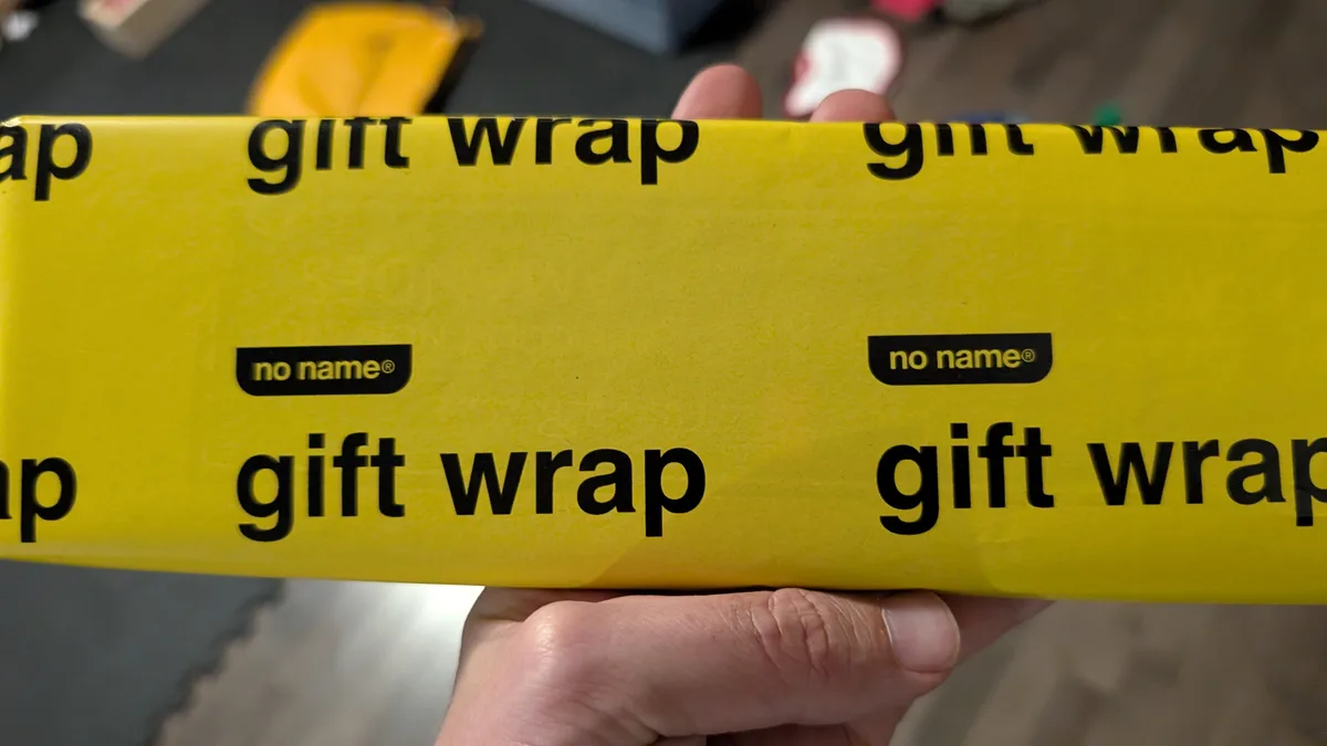 A hand holds a roll of bright yellow gift wrap with the words 'gift wrap' printed repeatedly in black. The brand name 'no…