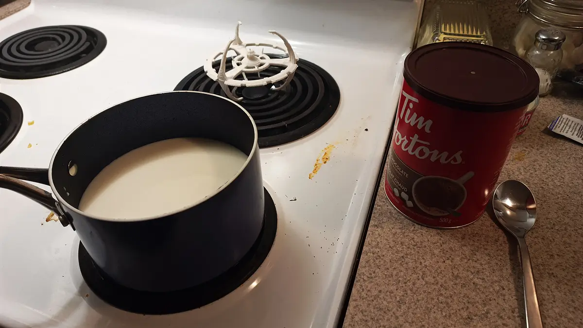 A black saucepan filled with milk sits on a white electric stove burner, next to a red Tim Hortons chocolate hot chocolate…