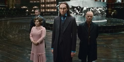 Four characters stand in a grand, dimly lit hall with a large fountain behind them. The central figure, a man in a dark…