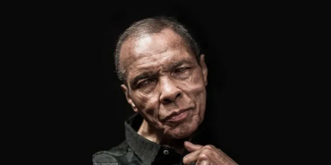 A close-up portrait of an older Black man with a serious, slightly skeptical expression, looking directly at the camera. He…