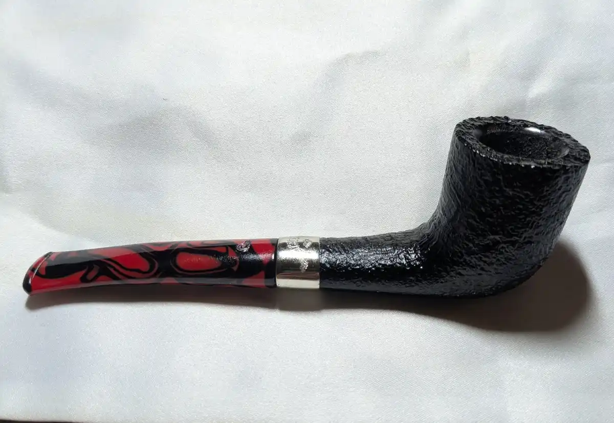 A smoking pipe with a black, textured bowl and a red-and-black marbled stem with a silver band, lying on a white fabric…