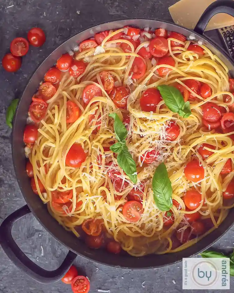 An overhead shot of a black skillet filled with spaghetti mixed with halved cherry tomatoes, grated cheese, and fresh basil…
