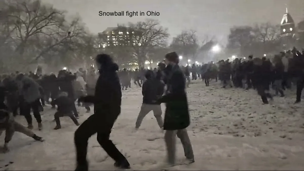 A blurry nighttime video shows a large crowd of people in winter coats running and throwing snowballs in a snowy park, with…