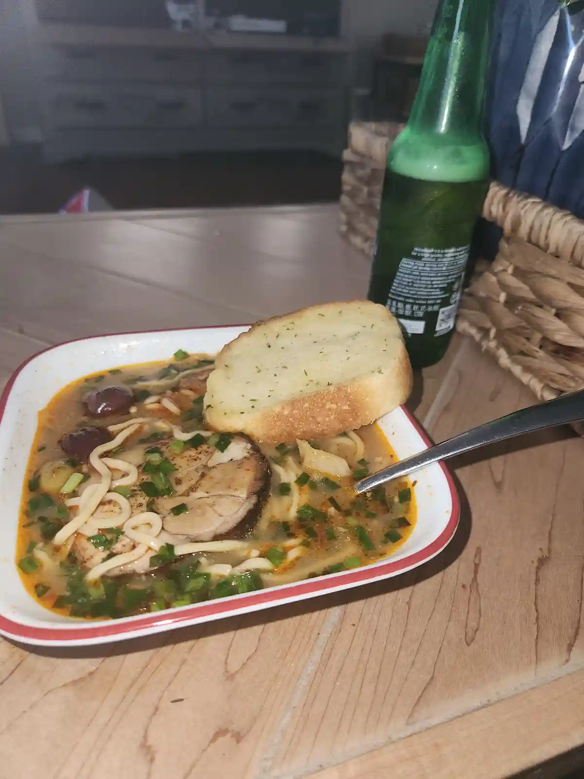 A close-up photo shows a bowl of noodle soup with meat, green onions, and a slice of garlic bread resting on the rim, next…