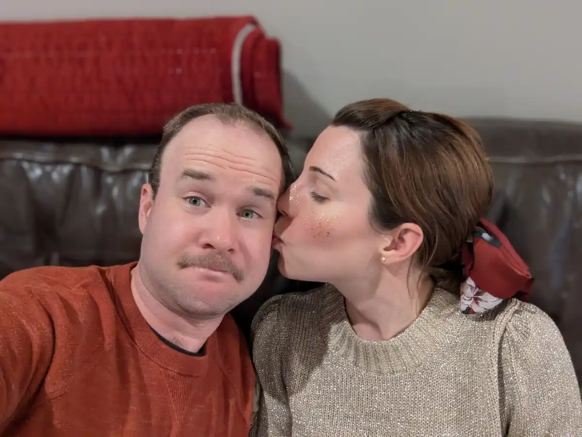 A man in an orange sweater sits on a couch looking bewildered as a woman in a sparkly gold sweater kisses his cheek. She has…