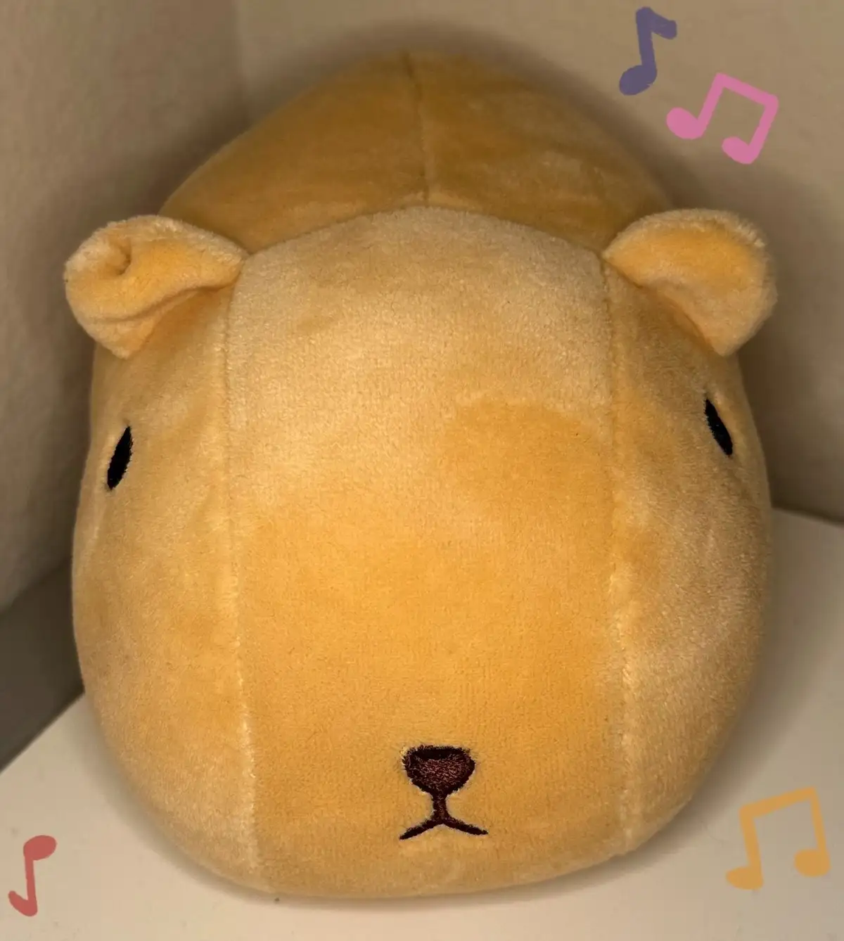 A close-up of a round, yellow plush toy resembling a capybara with simple black embroidered eyes and nose, sitting on a…