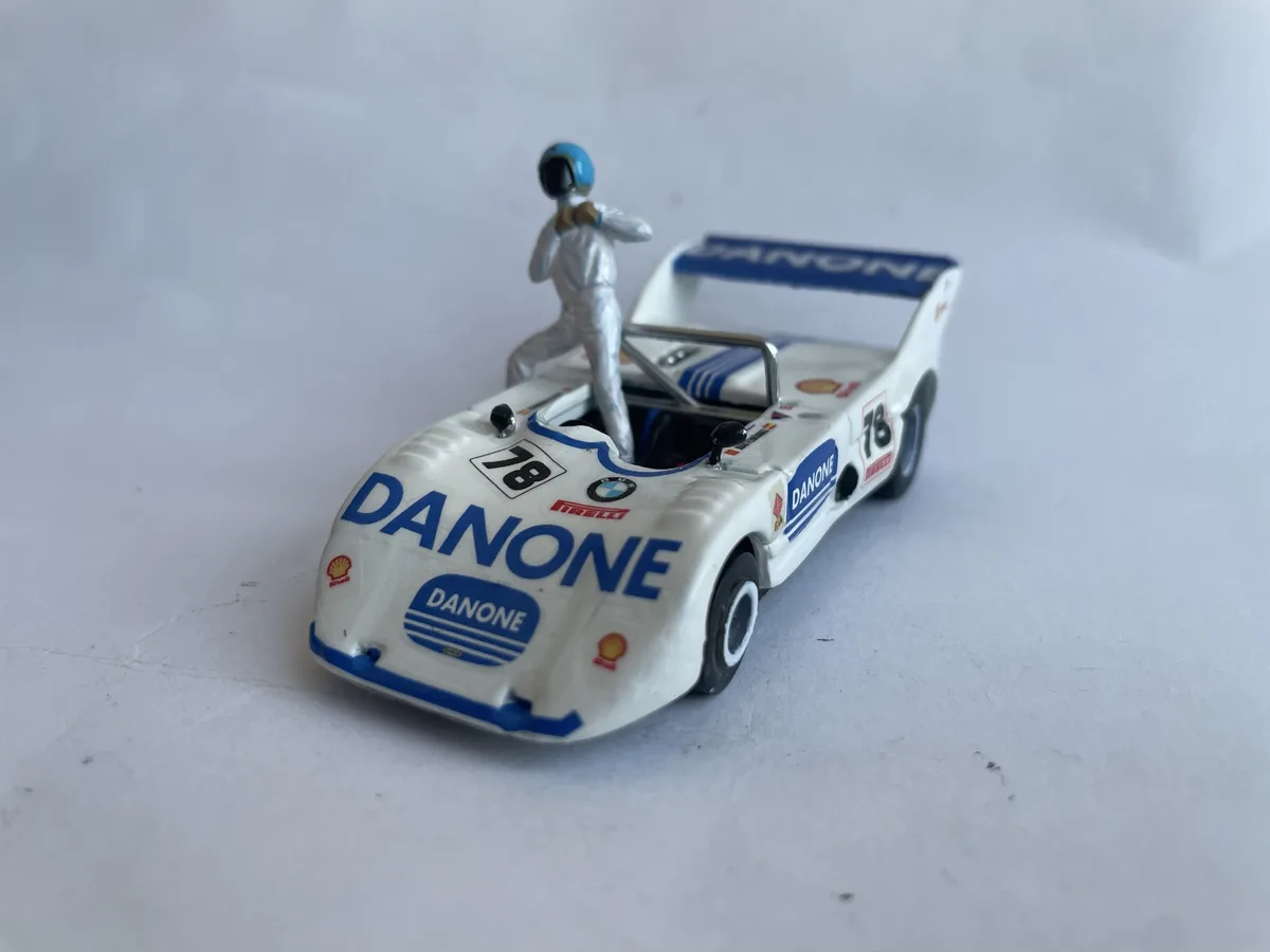A small white toy race car with blue and red sponsor logos, including 'DANONE' and 'PIRELLI', and the number 78, has a tiny…