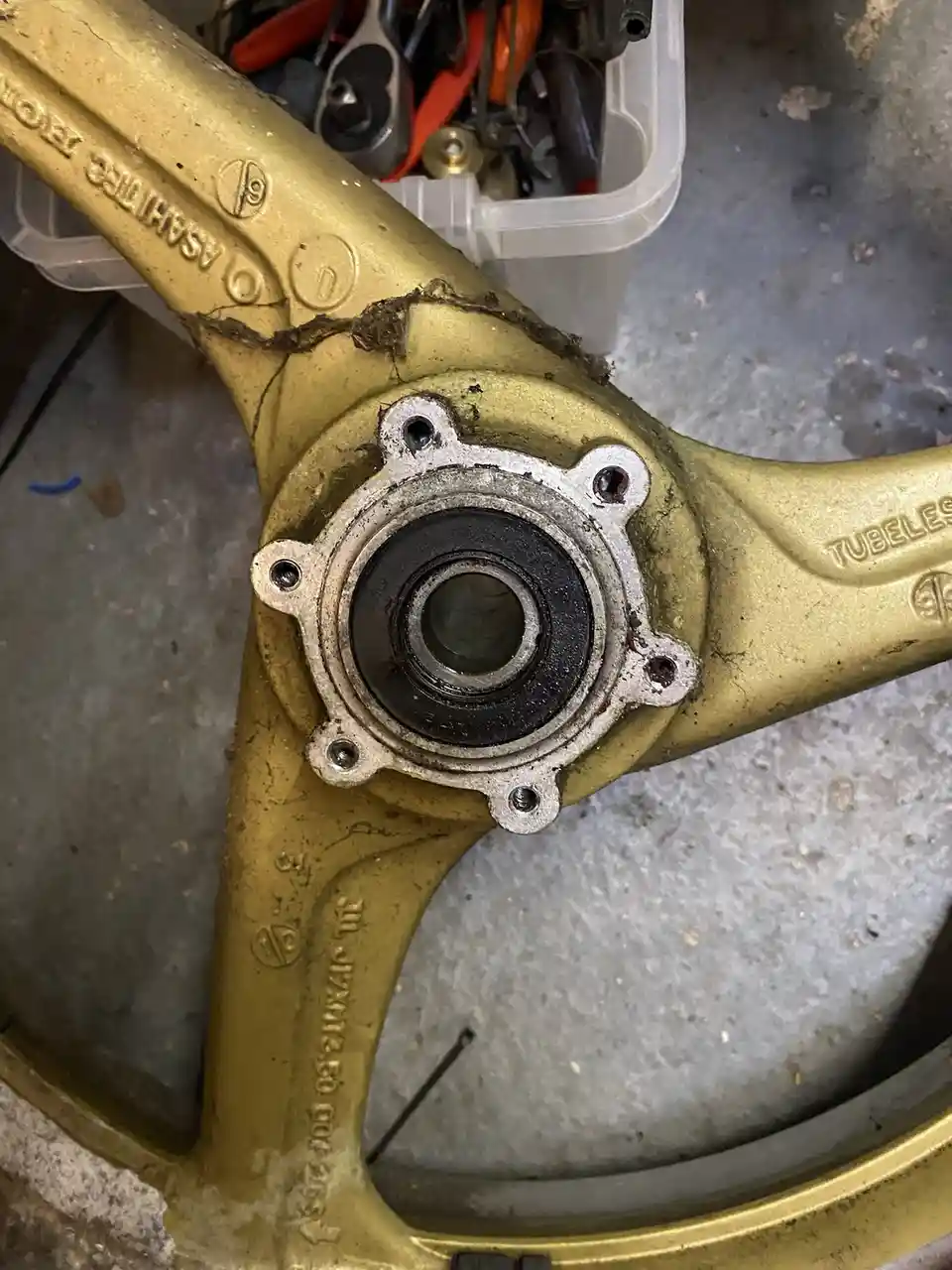 A close-up photo shows a gold-painted bicycle wheel hub with visible dirt and a cracked frame, set against a concrete floor…
