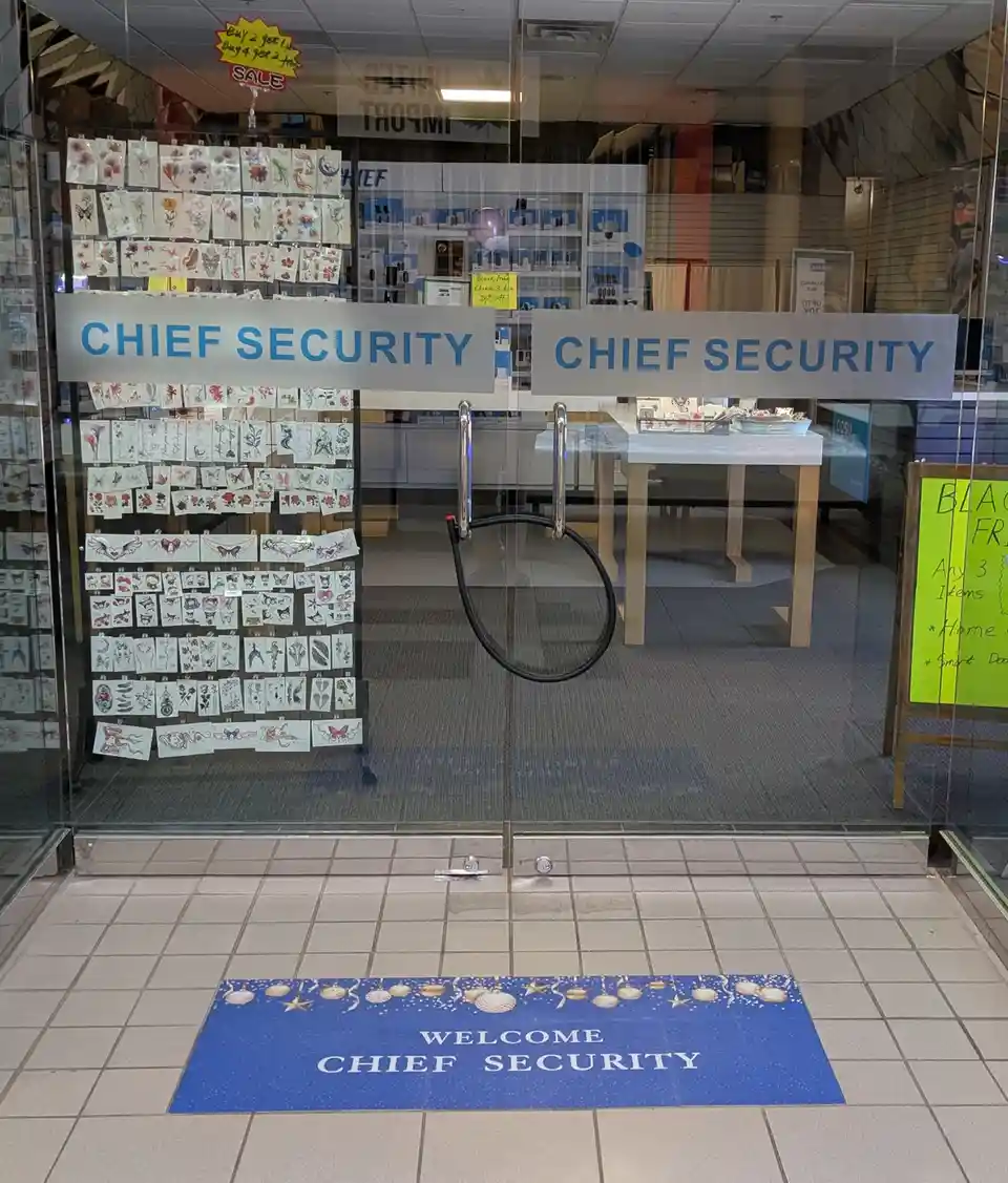 A store entrance with glass doors and a blue welcome mat that reads 'WELCOME CHIEF SECURITY'. Above the doors, two signs…