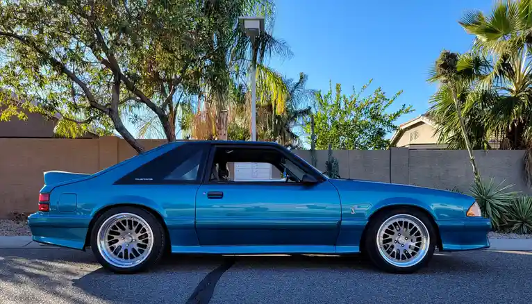 A side profile of a modified blue 1990s Ford Mustang S/C coupe parked on a residential street, featuring aftermarket silver…