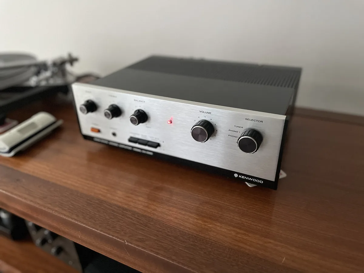 A close-up photo of a silver and black Kenwood stereo amplifier sitting on a wooden shelf, with a red power indicator light…