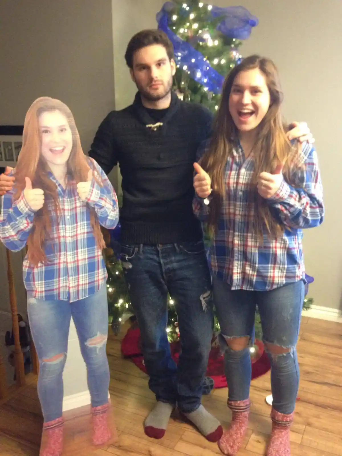 Three people stand in front of a decorated Christmas tree; a cardboard cutout of a smiling woman in a plaid shirt and ripped…