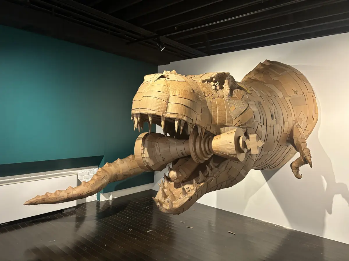 A large, detailed sculpture of a Tyrannosaurus Rex made entirely of cardboard is mounted in an art gallery, its mouth open…