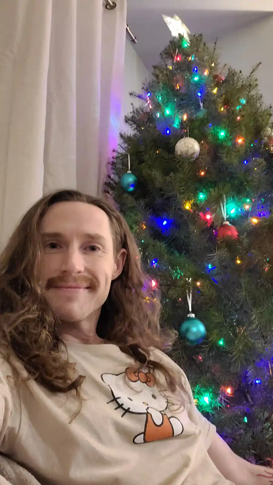 A man with long brown hair and a mustache smiles at the camera while wearing a beige t-shirt with a Hello Kitty graphic…