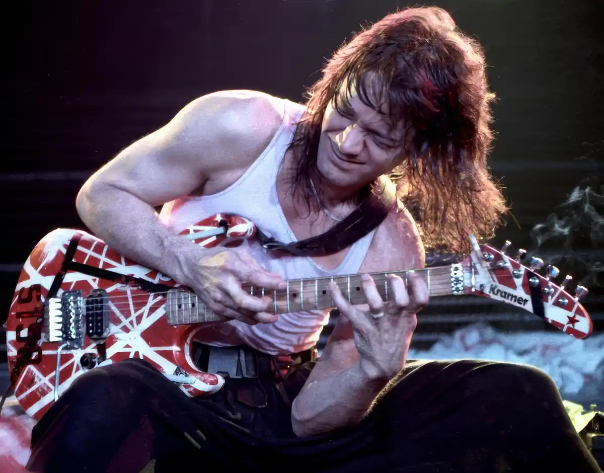 A man with long brown hair, wearing a white tank top and dark pants, is intensely playing a red and white electric guitar…