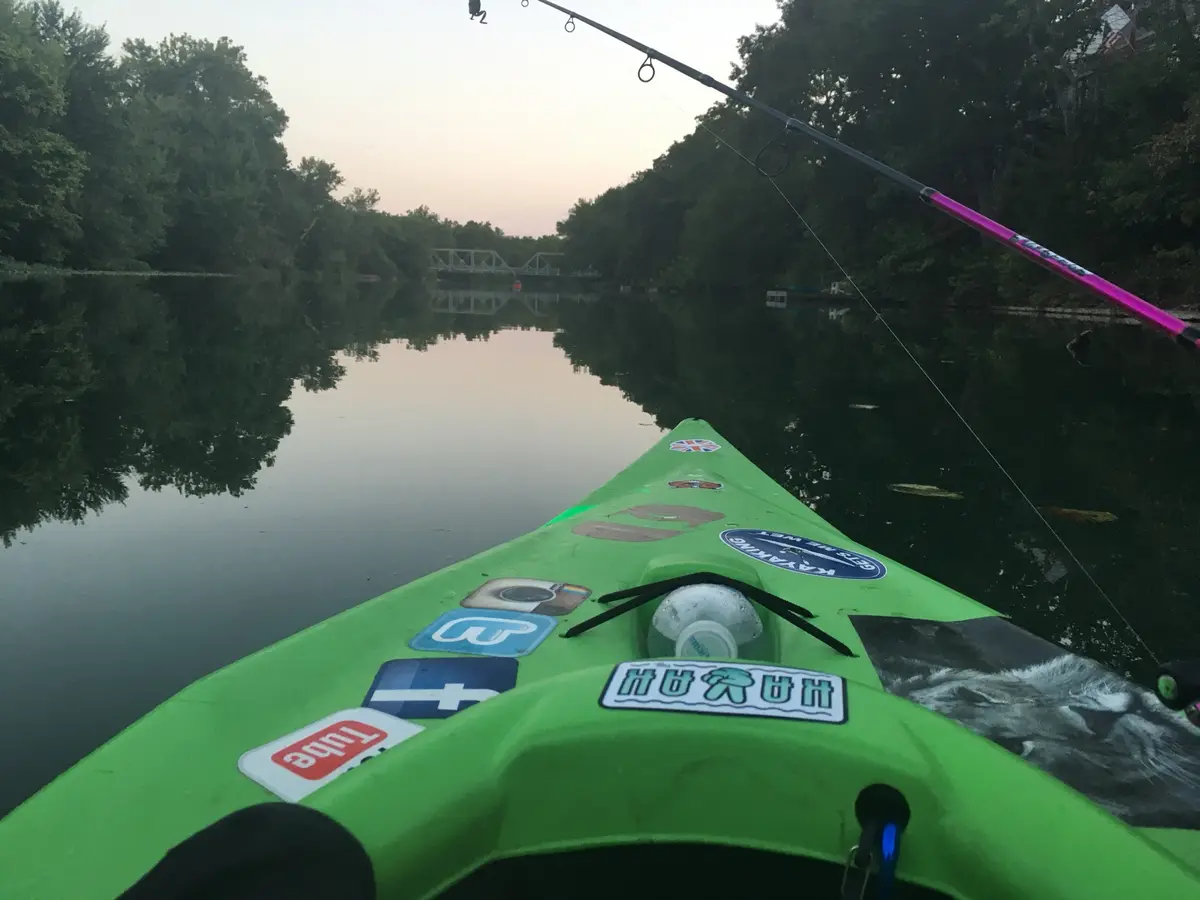 A first-person view from a bright green kayak on a calm river at dusk, with trees lining the banks and a distant bridge…