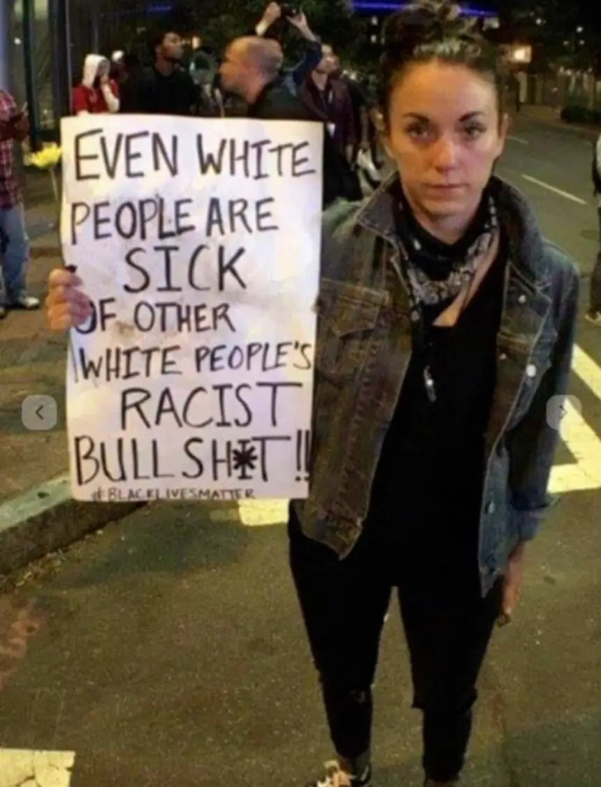 A woman stands outdoors at night holding a handmade sign that reads 'EVEN WHITE PEOPLE ARE SICK OF OTHER WHITE PEOPLE'S…