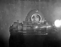A grainy, black-and-white image shows a person inside a vintage, cylindrical diving helmet with a circular viewport. The…