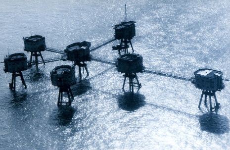 a blue-tinted aerial photograph shows several large, cylindrical, metal structures mounted on legs, arranged in a networked…