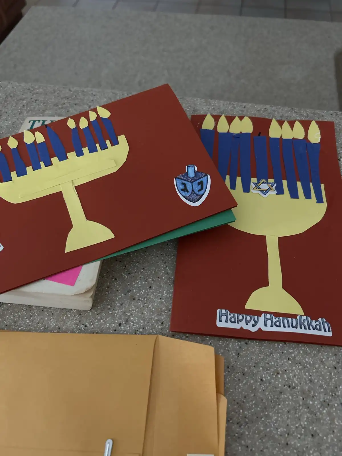 Two handmade Hanukkah greeting cards with paper cutouts of menorahs, blue candles, and Jewish symbols like a dreidel and…