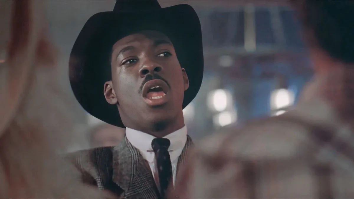 A man wearing a black cowboy hat, white collared shirt, and patterned suit jacket has his mouth open in an expression of…