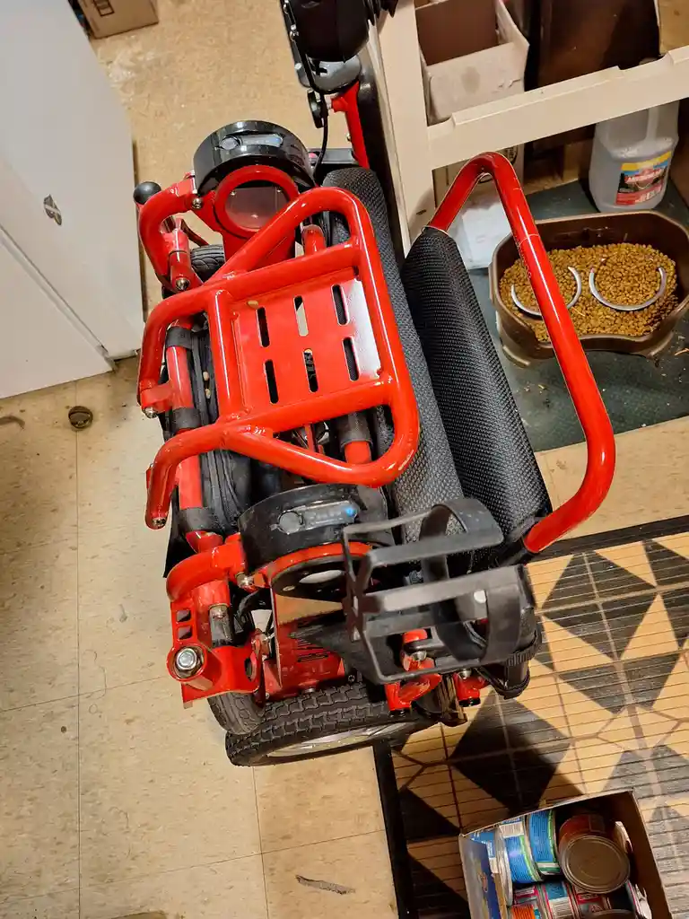 A red electric wheelchair is photographed from a high angle, showing its frame, wheels, and black seat. In the background, a…